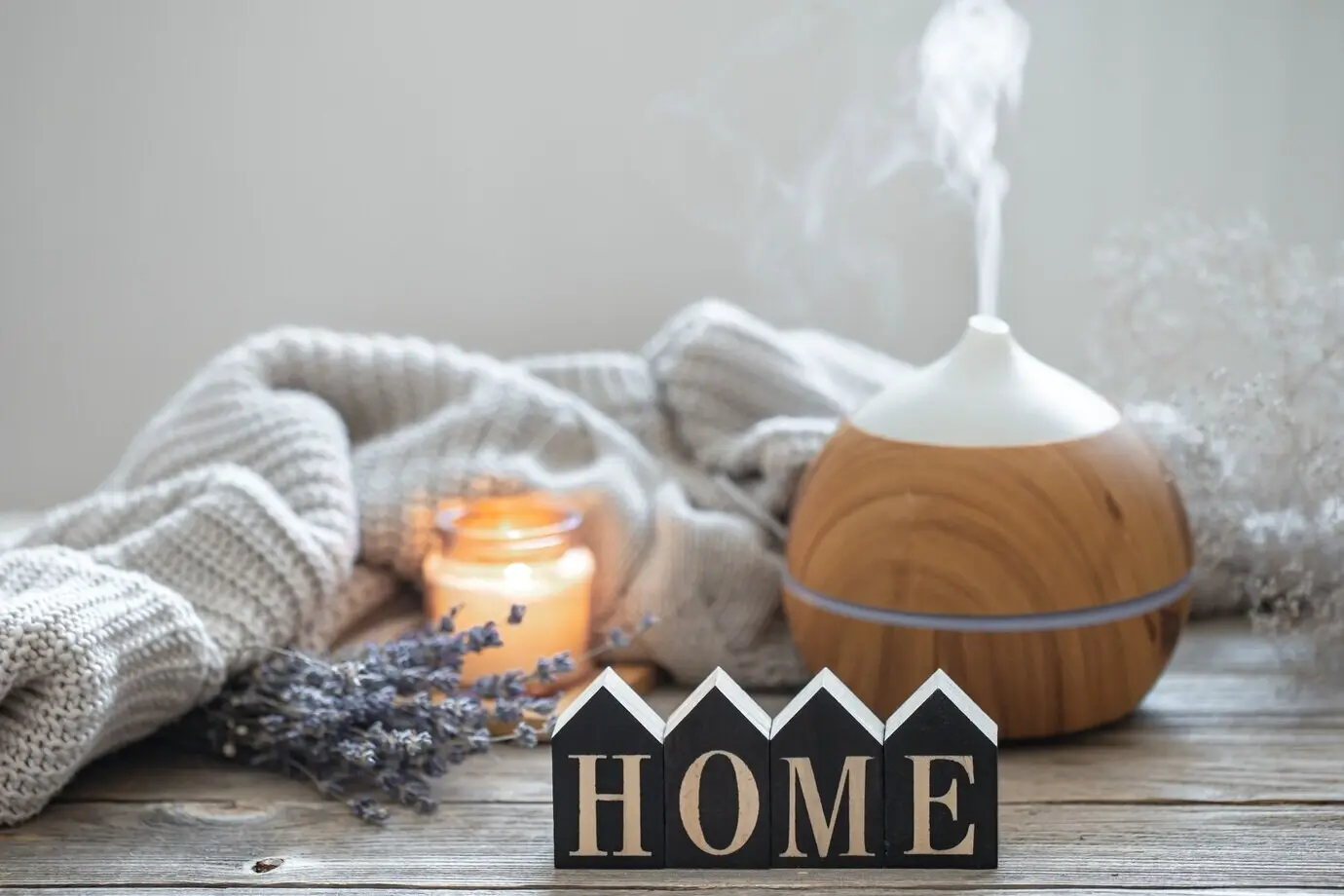 An aroma still life featuring a modern aroma oil diffuser on a wooden surface, with a knitted element, cozy details, and the decorative word “home.”