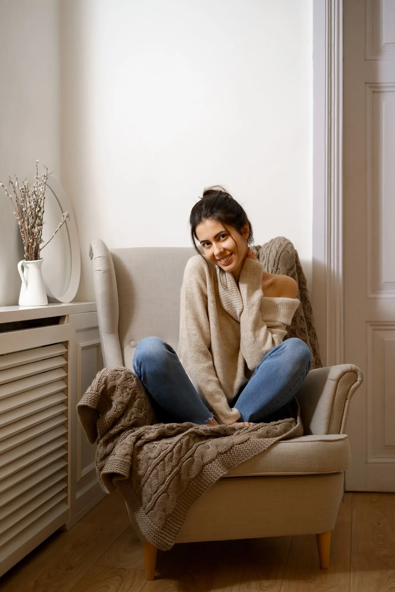 A smiling lady wearing smart, trendy attire is seated in an armchair indoors, in a relaxing atmosphere.