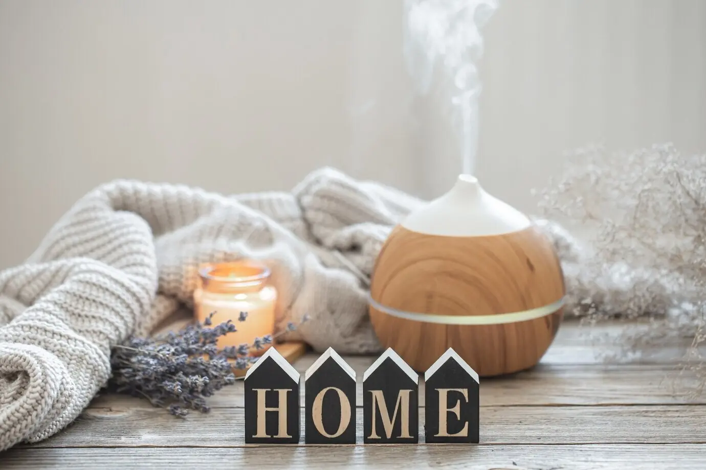 An aroma still life featuring a modern aroma oil diffuser on a wooden surface, with a knitted element, cozy details, and the decorative word “home.”